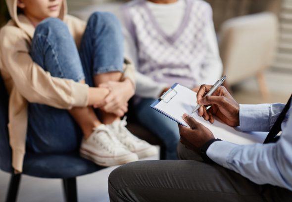 Primer plano de un psicólogo afroamericano tomando notas en un portapapeles en una sesión de terapia para niños.