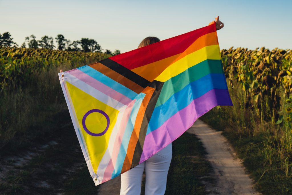 Una joven corre en el campo con la bandera Rainbow LGBTQ ondeando en el viento hecha de material de seda en el fondo del campo.