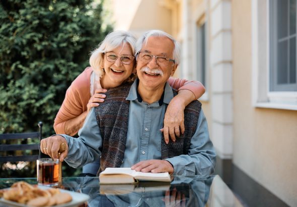 Mujer mayor feliz abrazando a su marido en el patio de la residencia de ancianos y mirando a cámara.